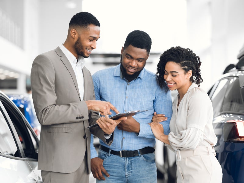 Automotive dealership representative showing information on a tablet to two customers beside a vehicle in a showroom.